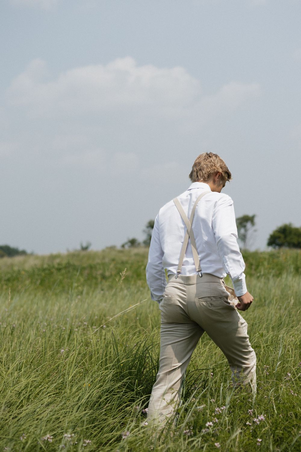 Person in white shirt and suspenders walking through tall grass field.