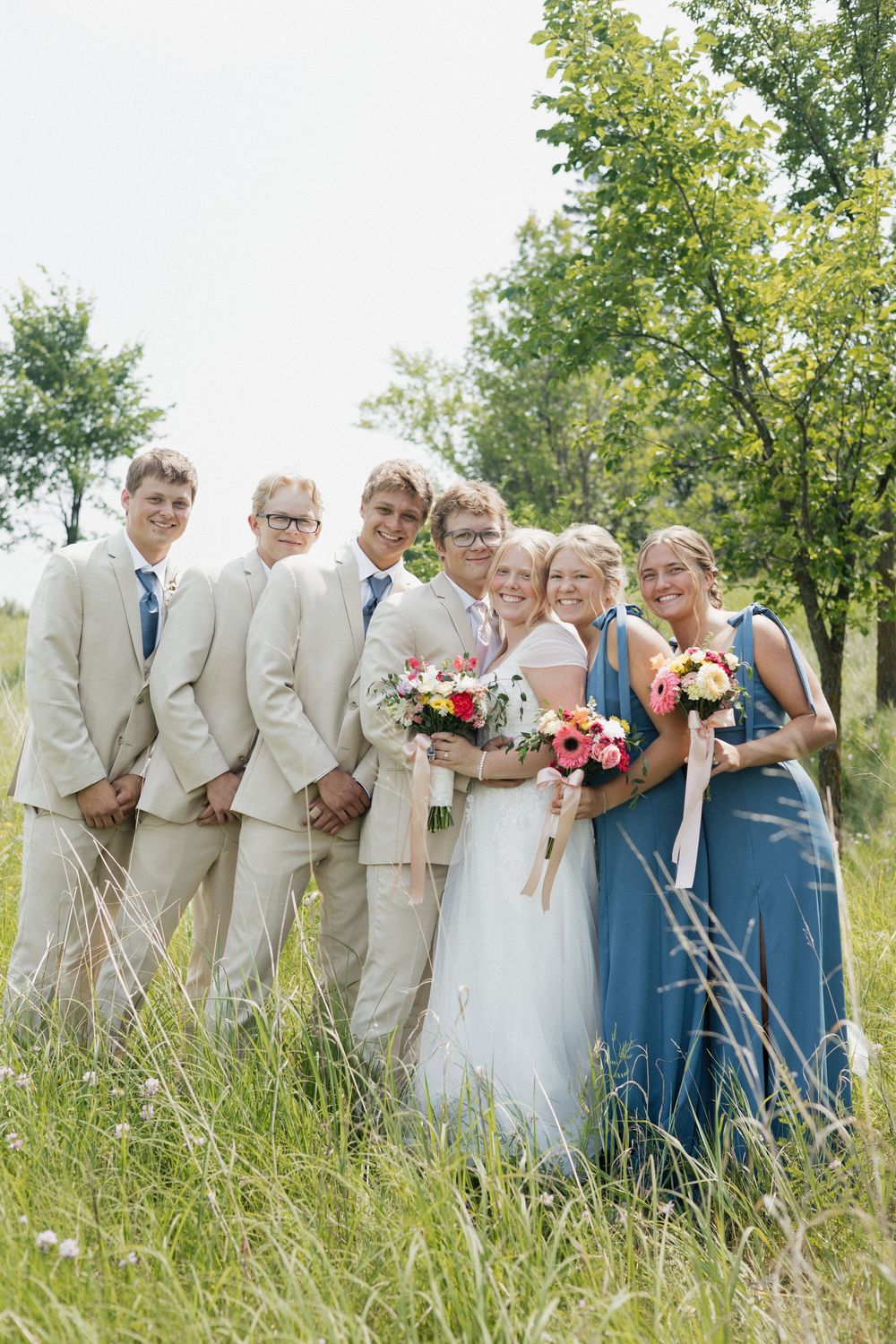 Wedding party poses together in tall grass wearing light beige suits and blue dresses.