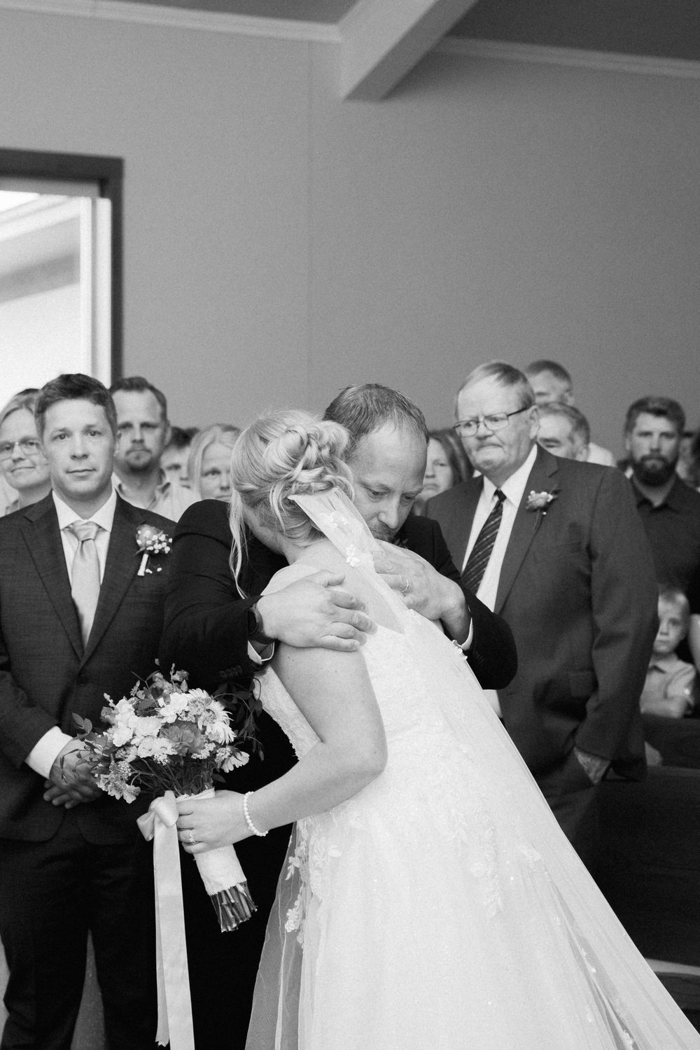 A married couple sharing their first kiss during their wedding ceremony while guests look on in black and white.