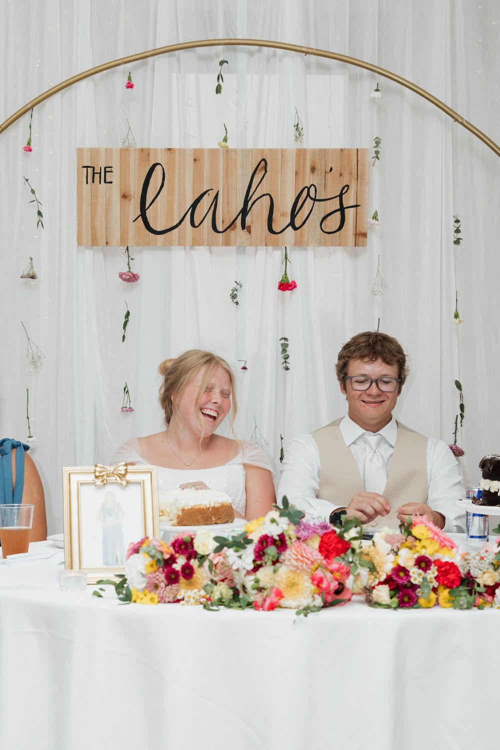 A newlywed couple sits at their reception table beneath a rustic wooden sign and floral decorations.