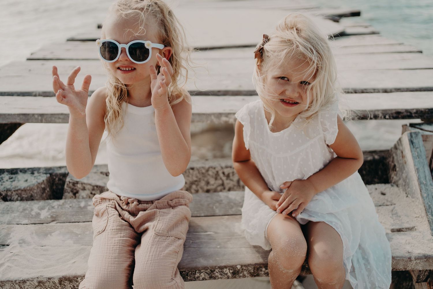 Two children sit on wooden steps by the water, one wearing white sunglasses and making a peace sign. Puerto Morelos Family Photography.