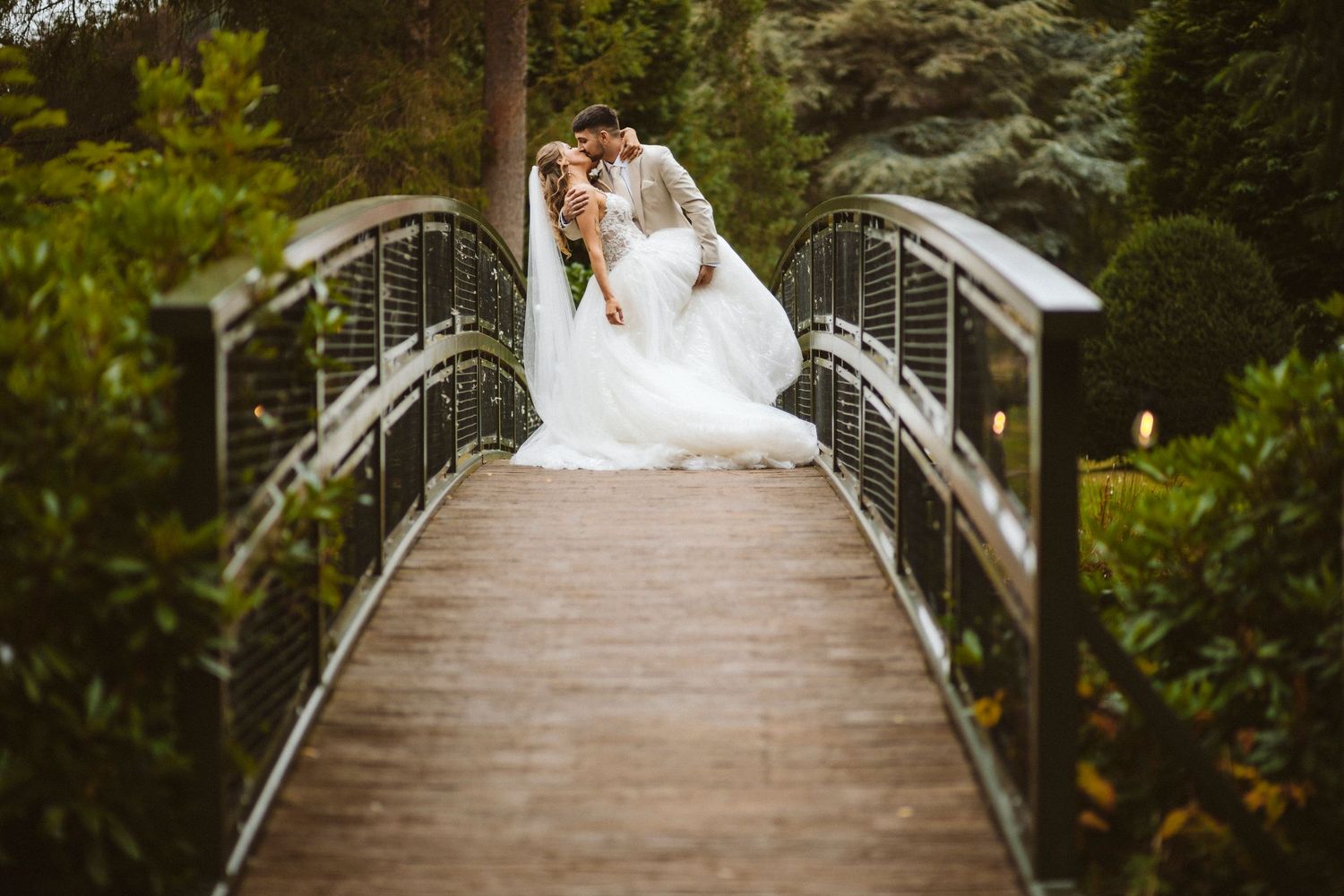 A couple sits together on a curved wooden bridge surrounded by lush greenery at their wedding celebration.