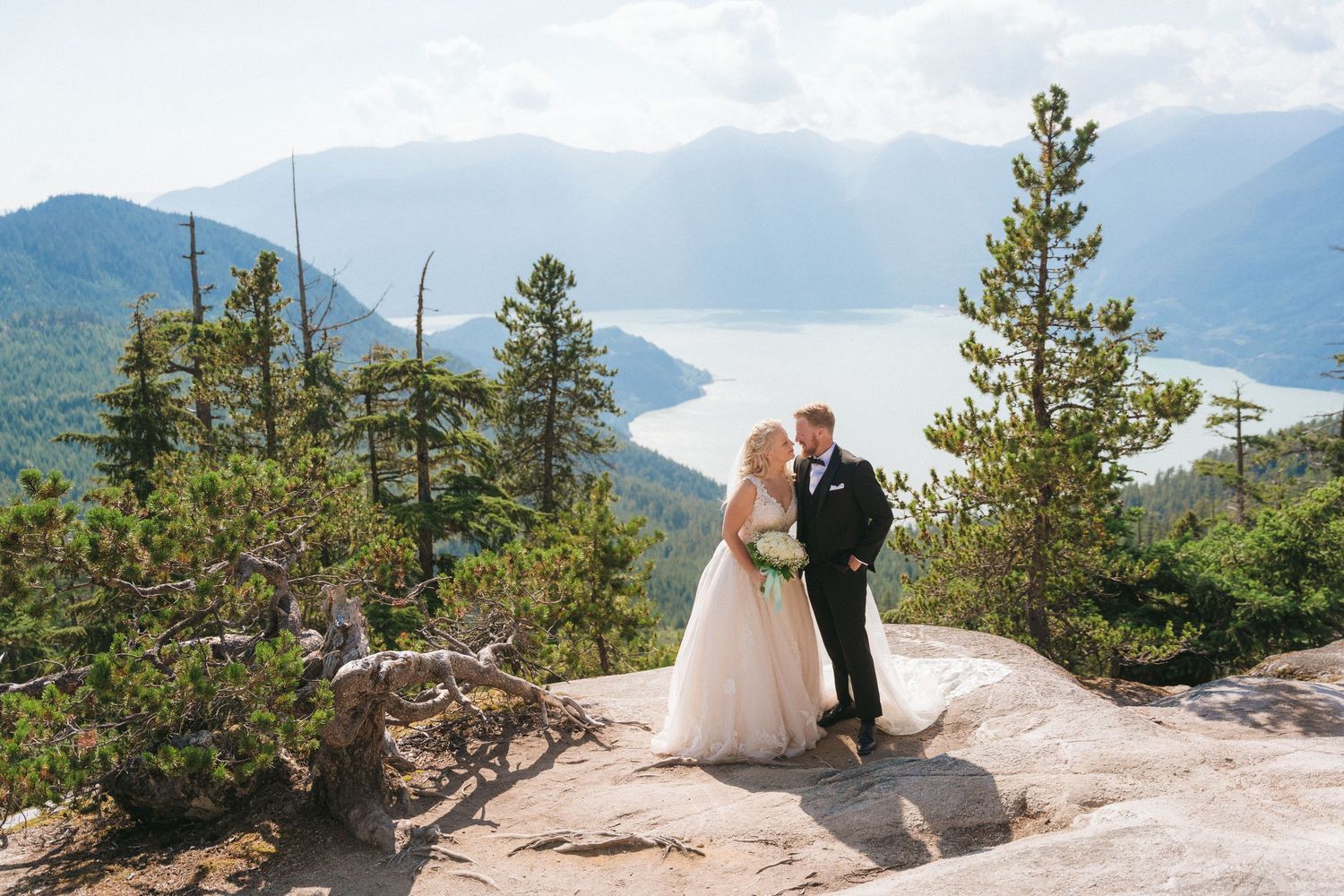 A wedding couple stands on a rocky overlook with mountain and lake views.