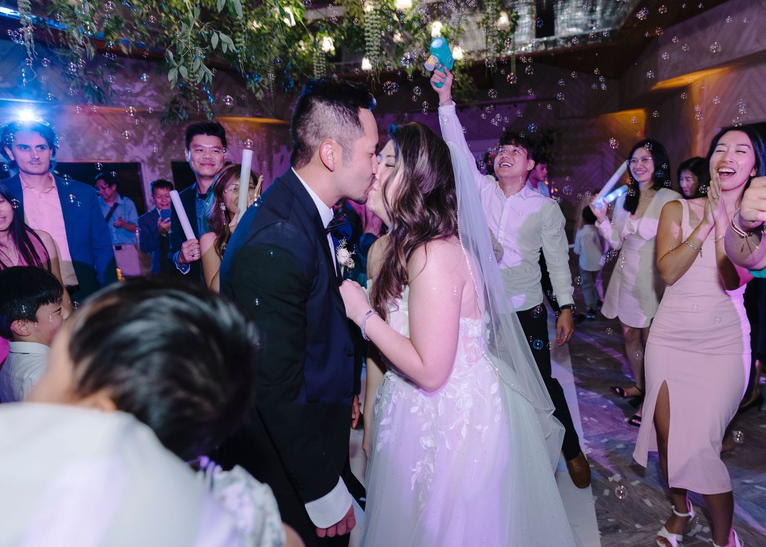 Wedding reception guests dance and celebrate on a crowded dance floor with colorful lighting effects.