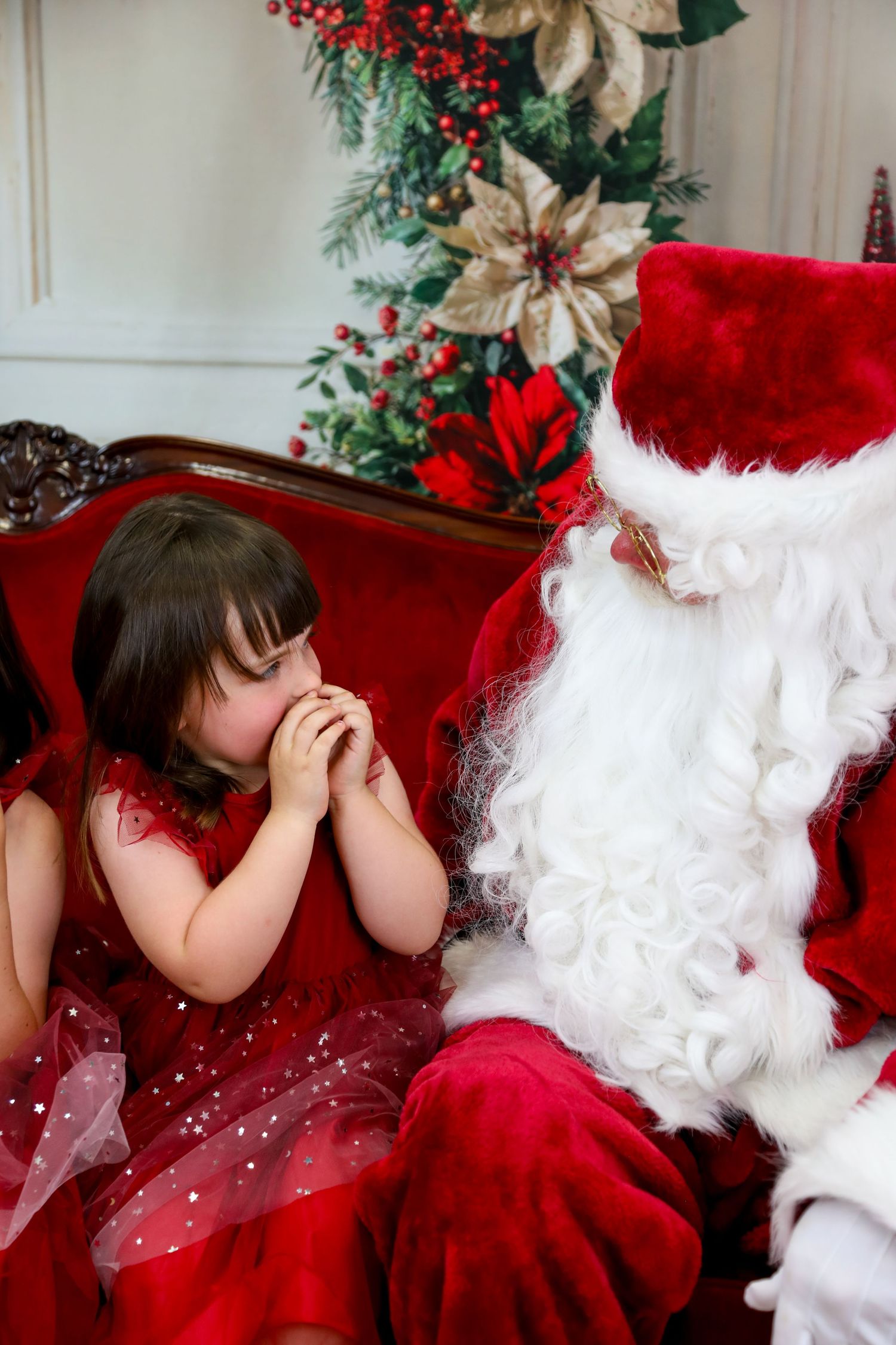A small child in a red dress laughing while sitting next to someone in a traditional holiday costume on a red couch.