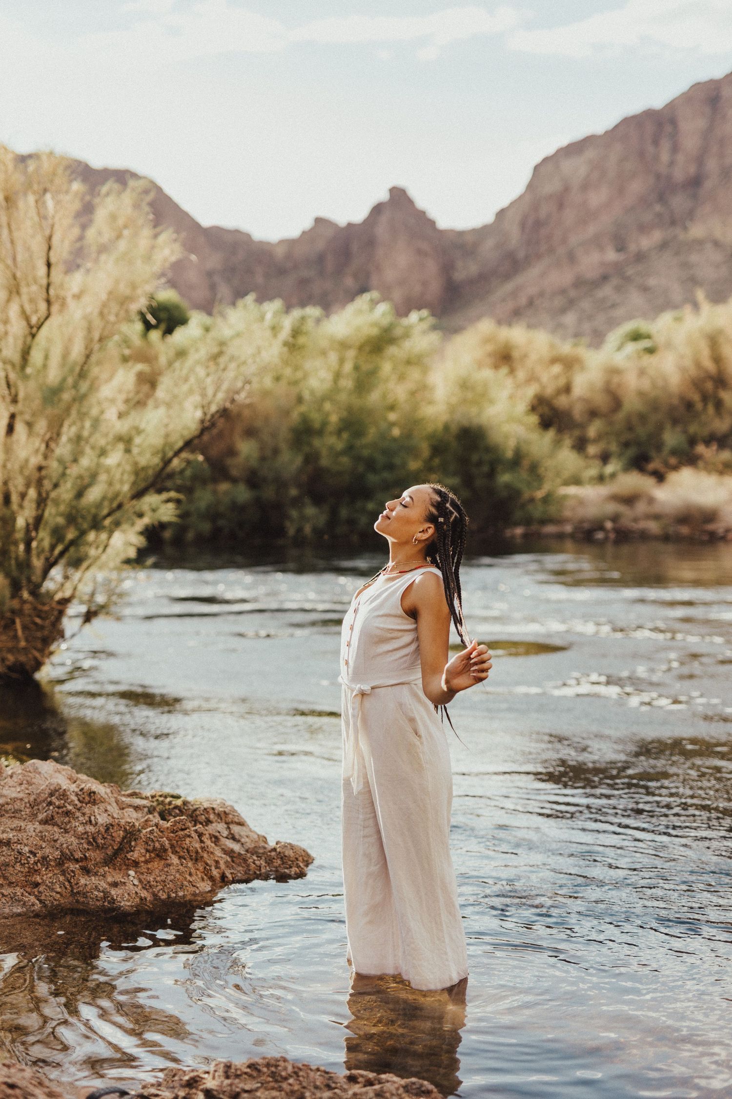 Person in flowy white dress stands peacefully in flowing river with rugged mountain peaks in background.