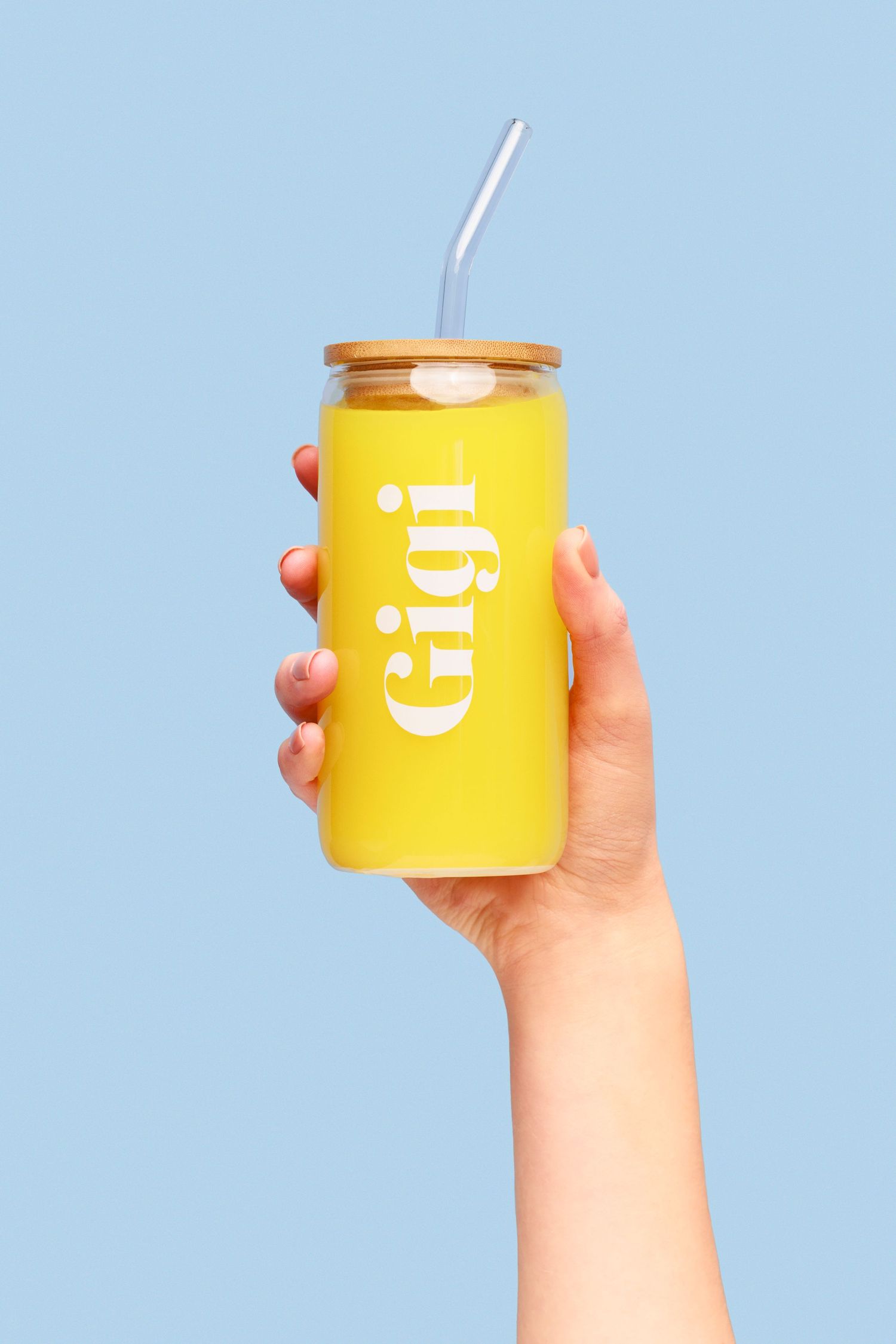Hand holds bright yellow drink in a Gigi branded tumbler against light blue background.