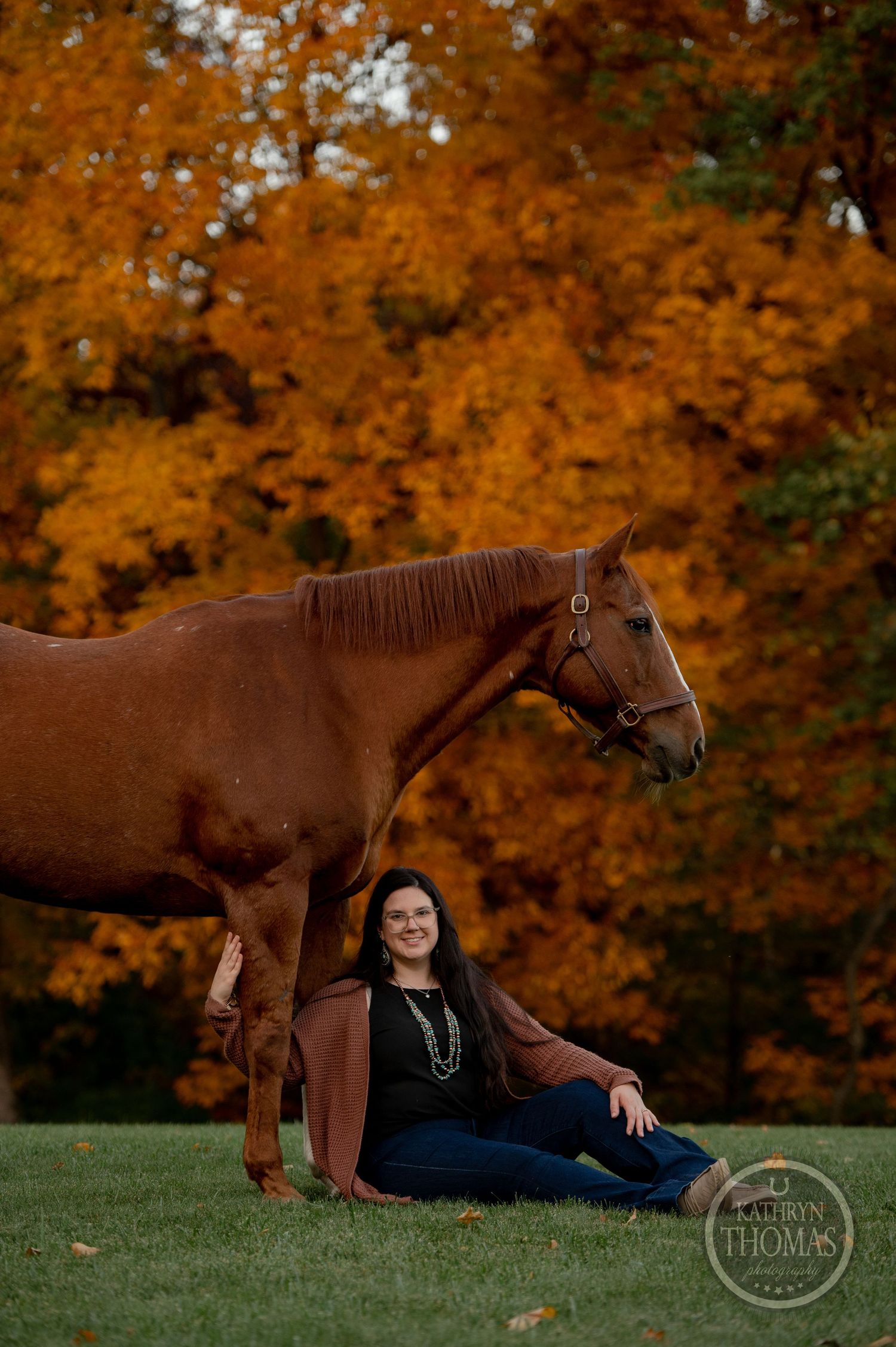 A beautiful brown horse poses beside a seated companion against a backdrop of bright orange fall trees.