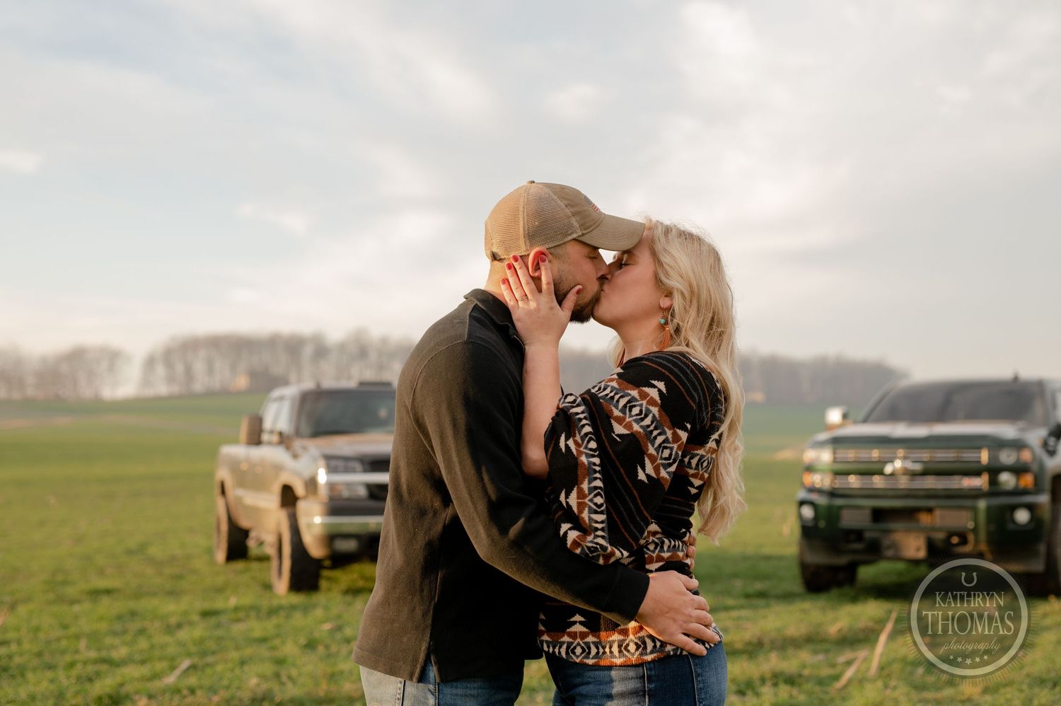 A couple shares a tender kiss in a field with trucks visible in the background.