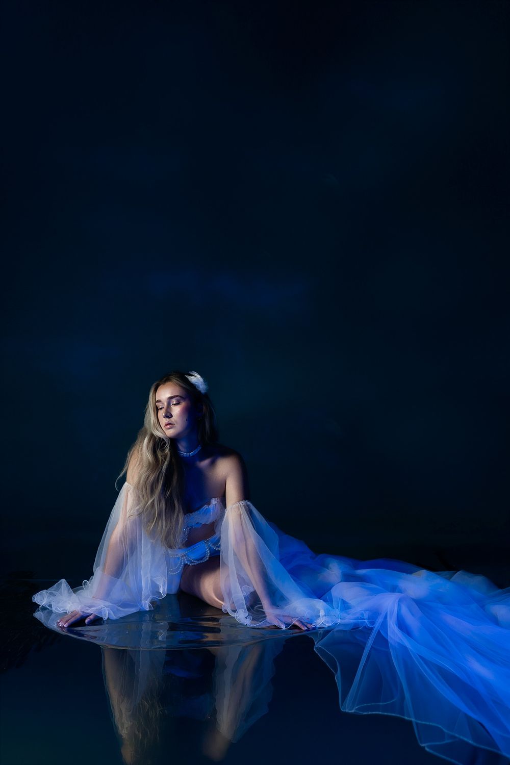 A woman in a flowing blue sheer gown sits gracefully on a reflective surface against a dark blue background.