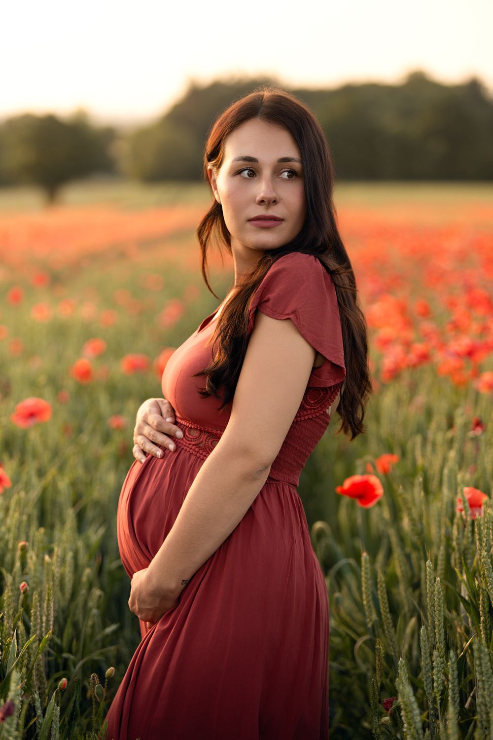 Pregnant woman in red dress cradling her baby bump in a vibrant poppy field at golden hour.