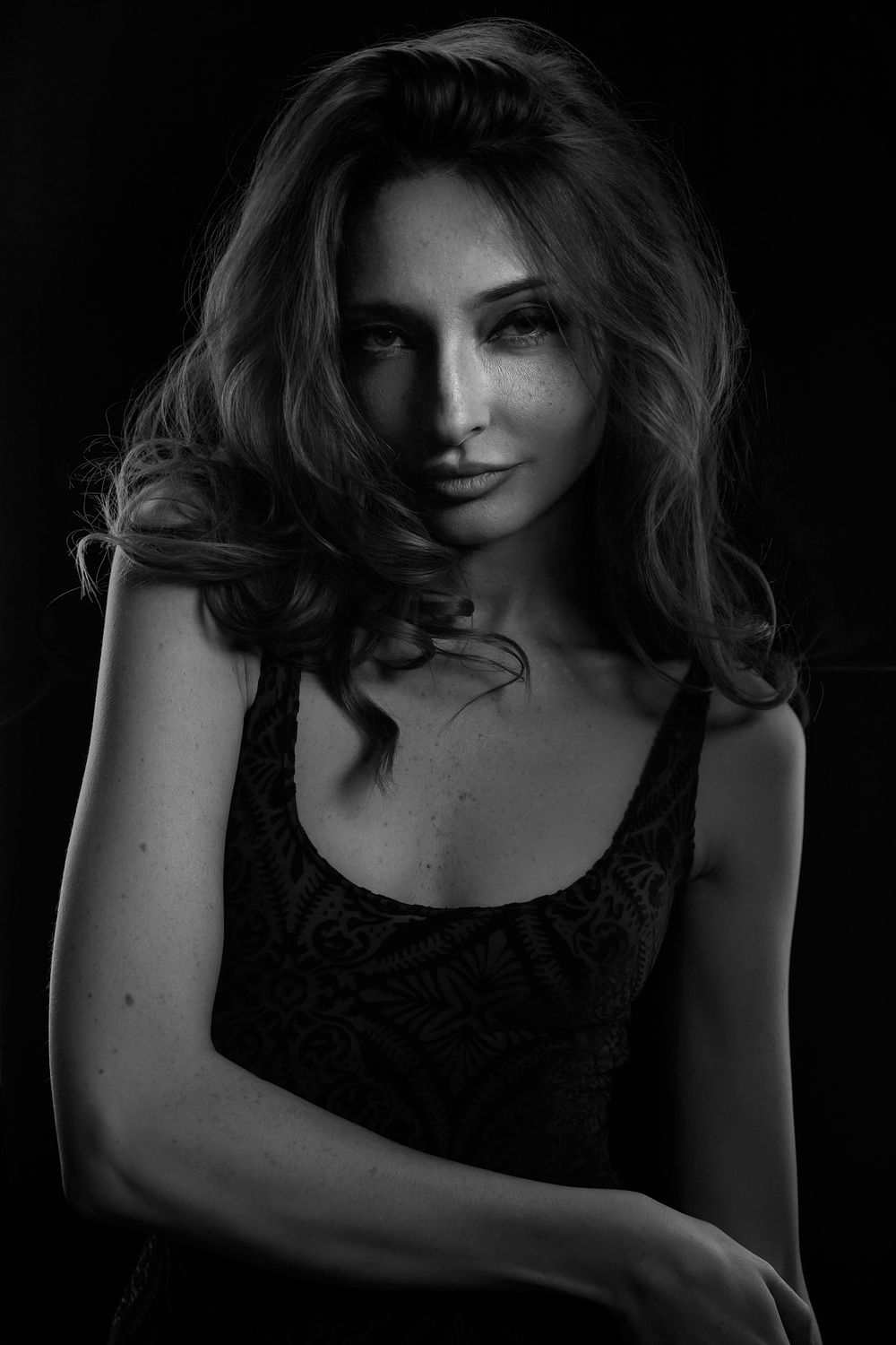 Moody black and white portrait of a young woman with wavy hair and freckles against dark background.