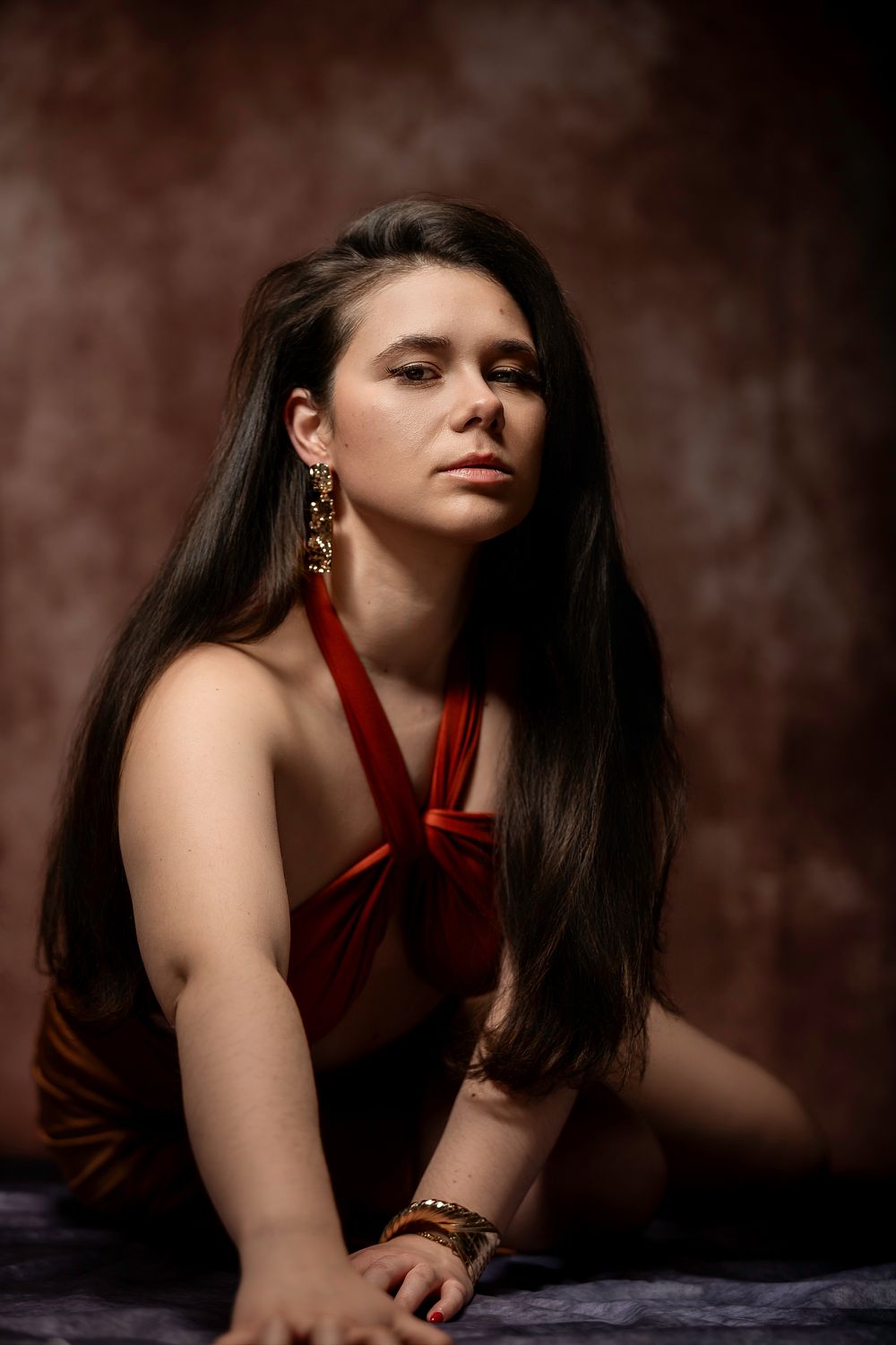 Young woman in red halter top with gold jewelry poses gracefully against a dark brown textured studio background.