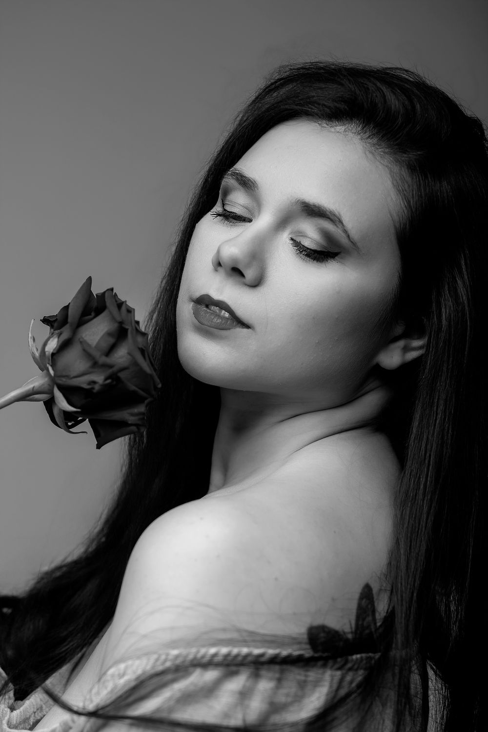 Black and white portrait of a young woman with closed eyes holding a rose near her shoulder.