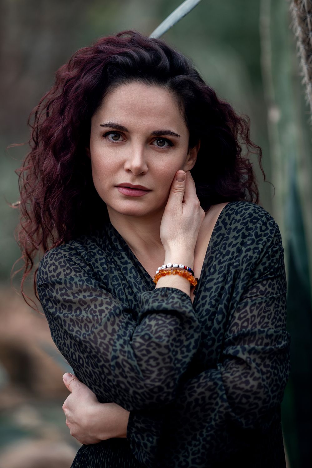A woman with curly dark red hair in a leopard print top, wearing an orange bracelet, posing outdoors.