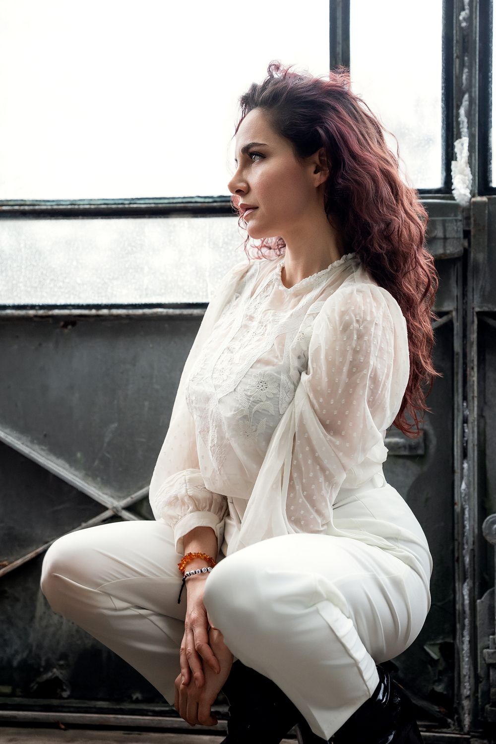 A woman with red wavy hair sits gracefully in white lace blouse and pants, gazing thoughtfully out an industrial window.