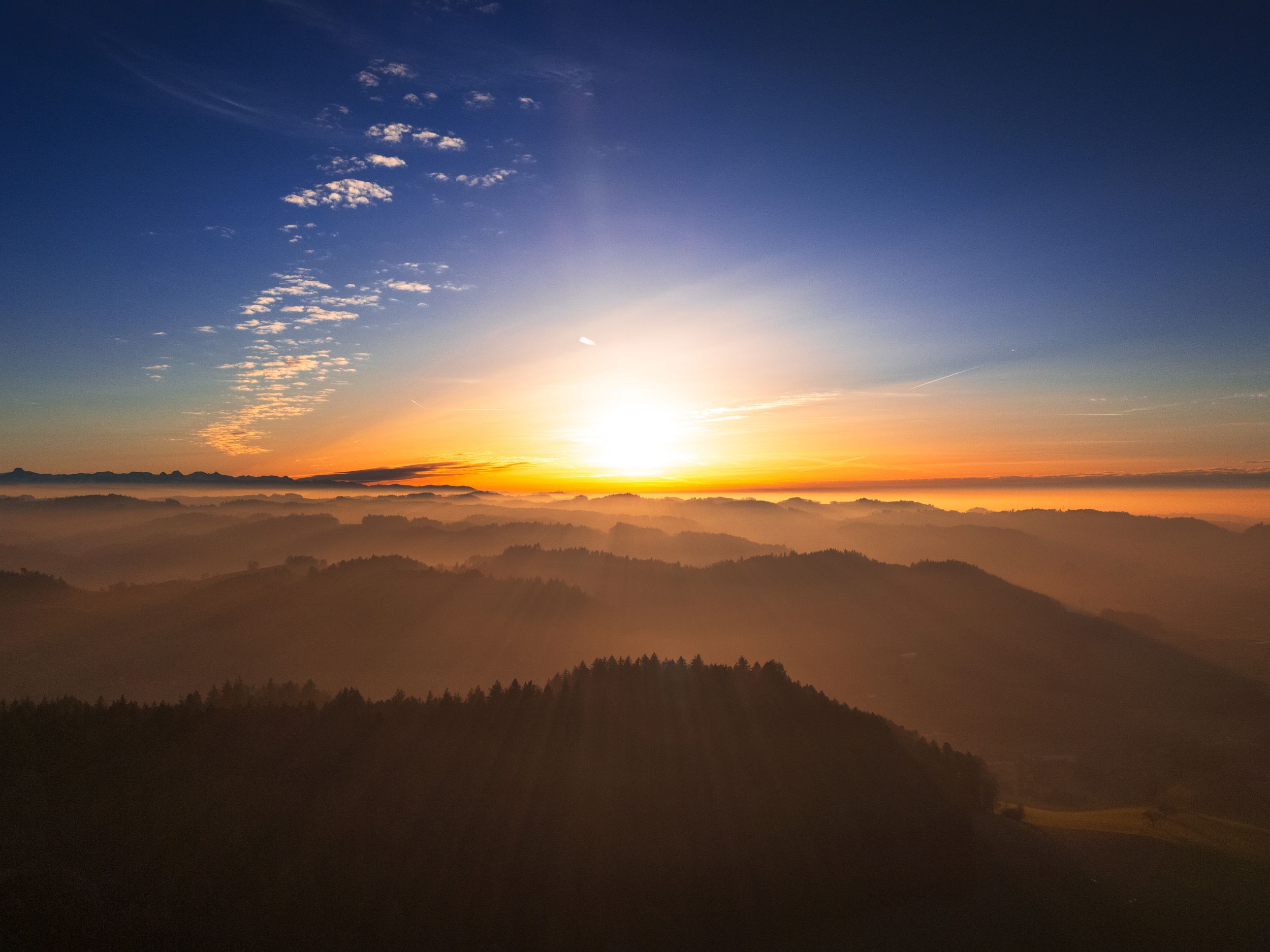 Golden sunset over misty mountain ranges with dramatic clouds and silhouetted forest.