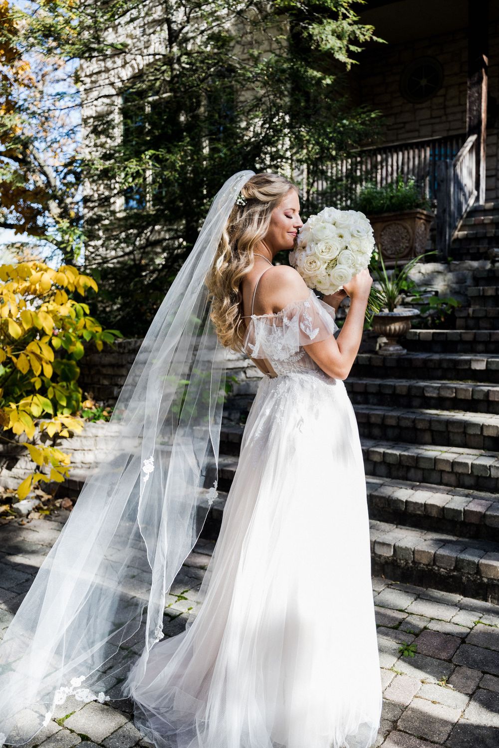 A bride in an elegant off-shoulder gown with long veil holds a white bouquet on stone steps.