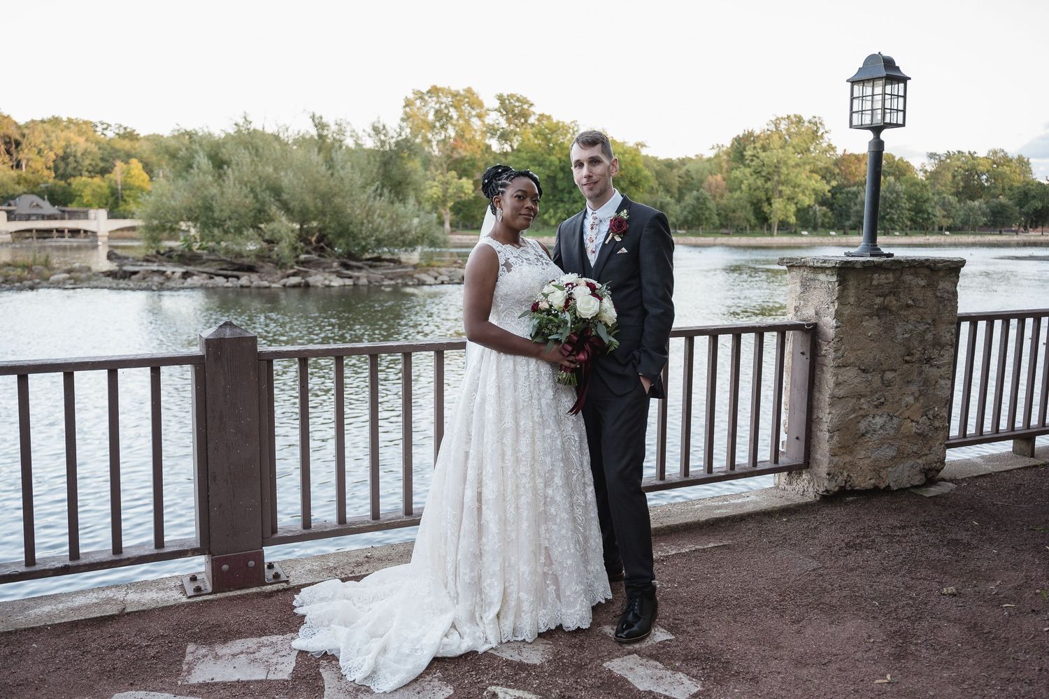 A couple poses for wedding photos on a bridge overlooking a serene lake at sunset.