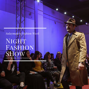Fashion model walking runway in trench coat and hat during night fashion show with blue lighting and audience.