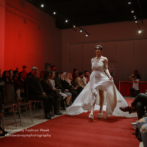 A model walks the runway in a flowing white wedding gown with a high slit during a fashion show with red backdrop lighting.