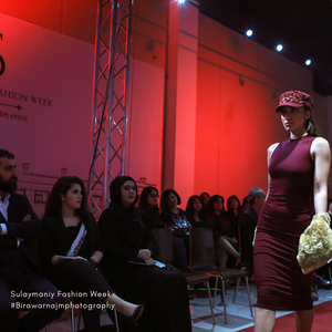 Model in burgundy fitted dress walks runway with red-lit wall backdrop during fashion week event.
