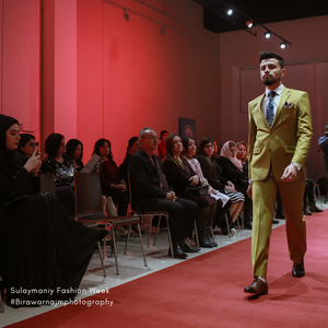 A model walks the runway in a mustard yellow suit during a fashion show with red walls and seated audience members.