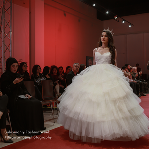 A model in a voluminous white tulle ball gown walks the runway at a fashion show against a red backdrop.