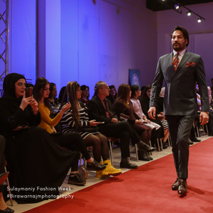 A model walks the runway in a gray suit with red tie during a fashion show as audience members look on.