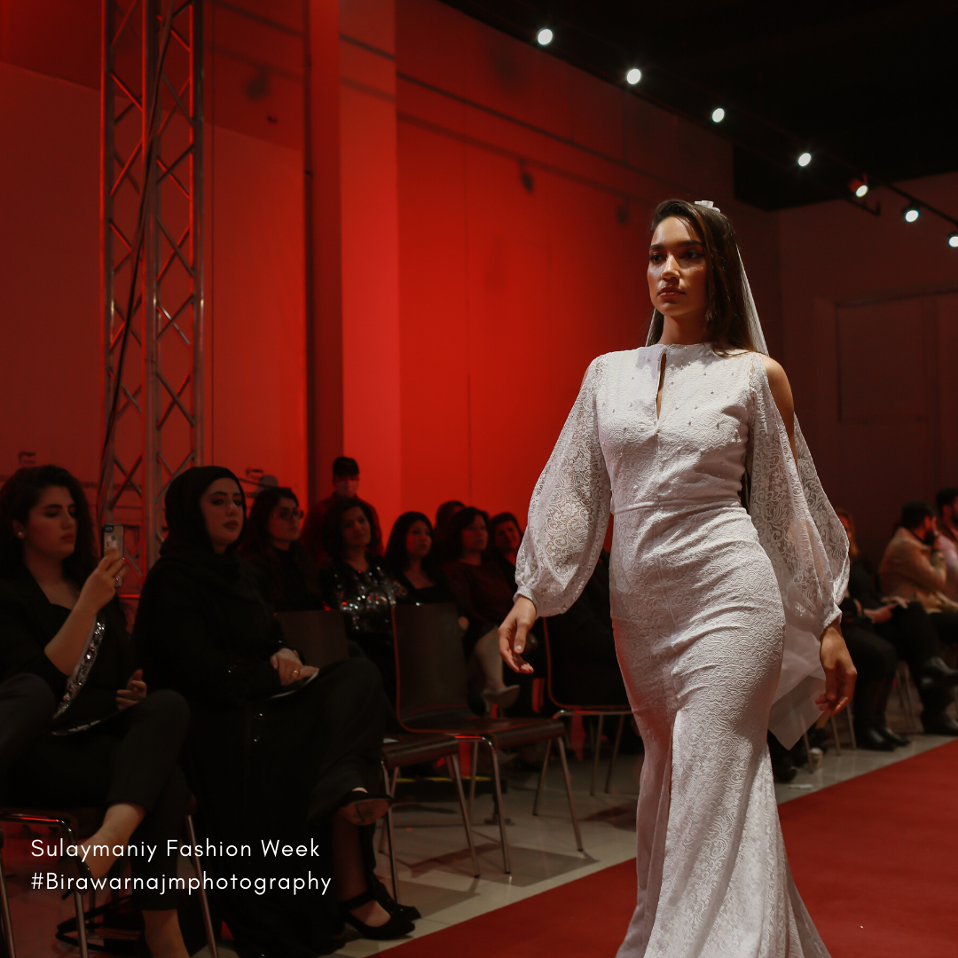 Elegant white wedding gown with voluminous sleeves on display at fashion show against vibrant red backdrop.