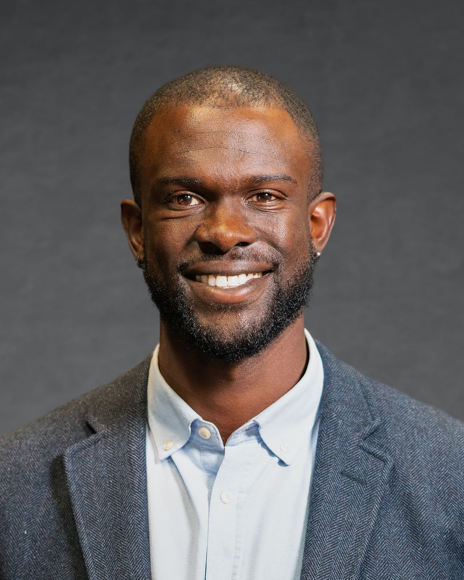 Professional headshot of a person wearing a gray blazer and white collared shirt against dark background.