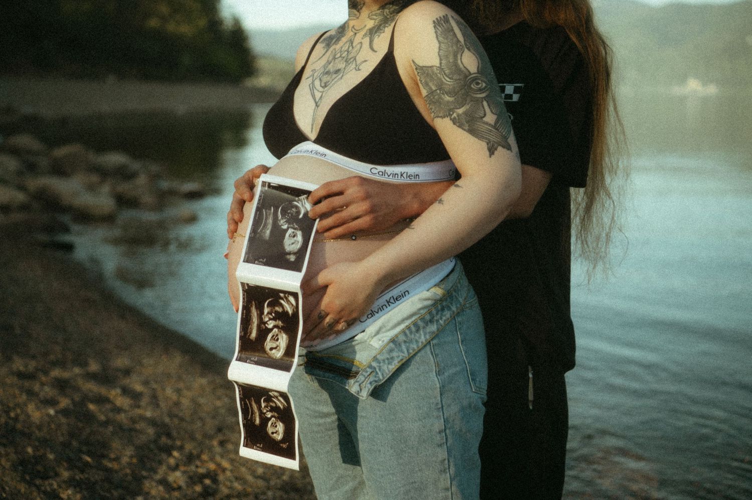 Pregnant person holding ultrasound photos against their belly while standing by a lake at sunset.