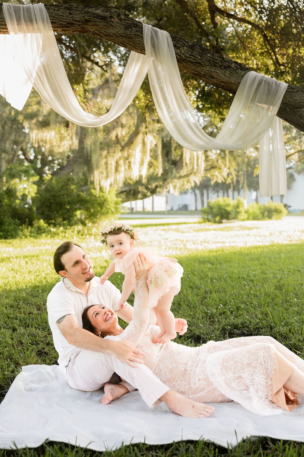 A family relaxes on a white blanket under a draped oak tree, with parents lovingly holding their baby girl outdoors.
