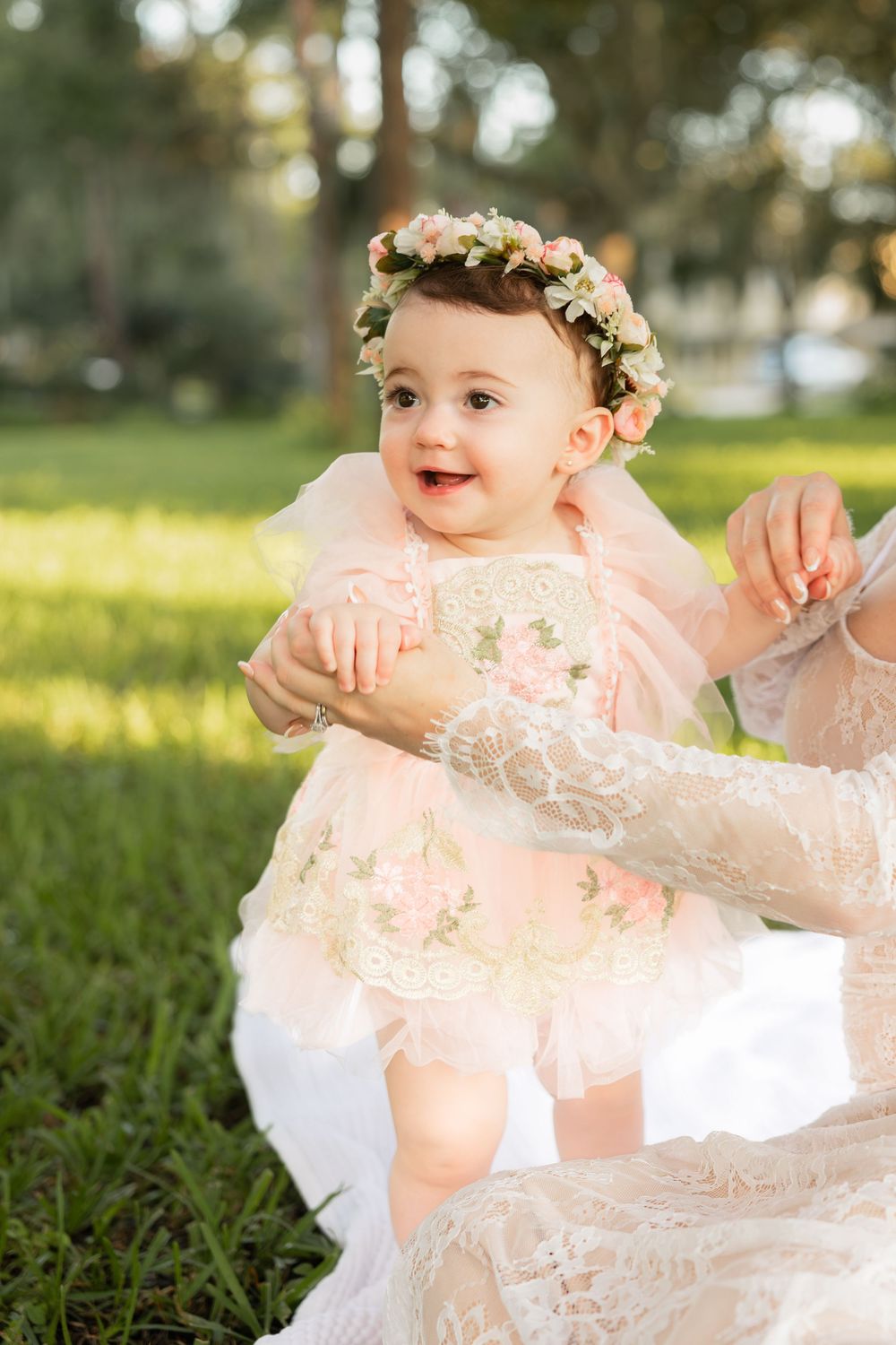 Happy baby girl wearing a floral crown and pink dress, smiling while sitting on grass in sunlight.