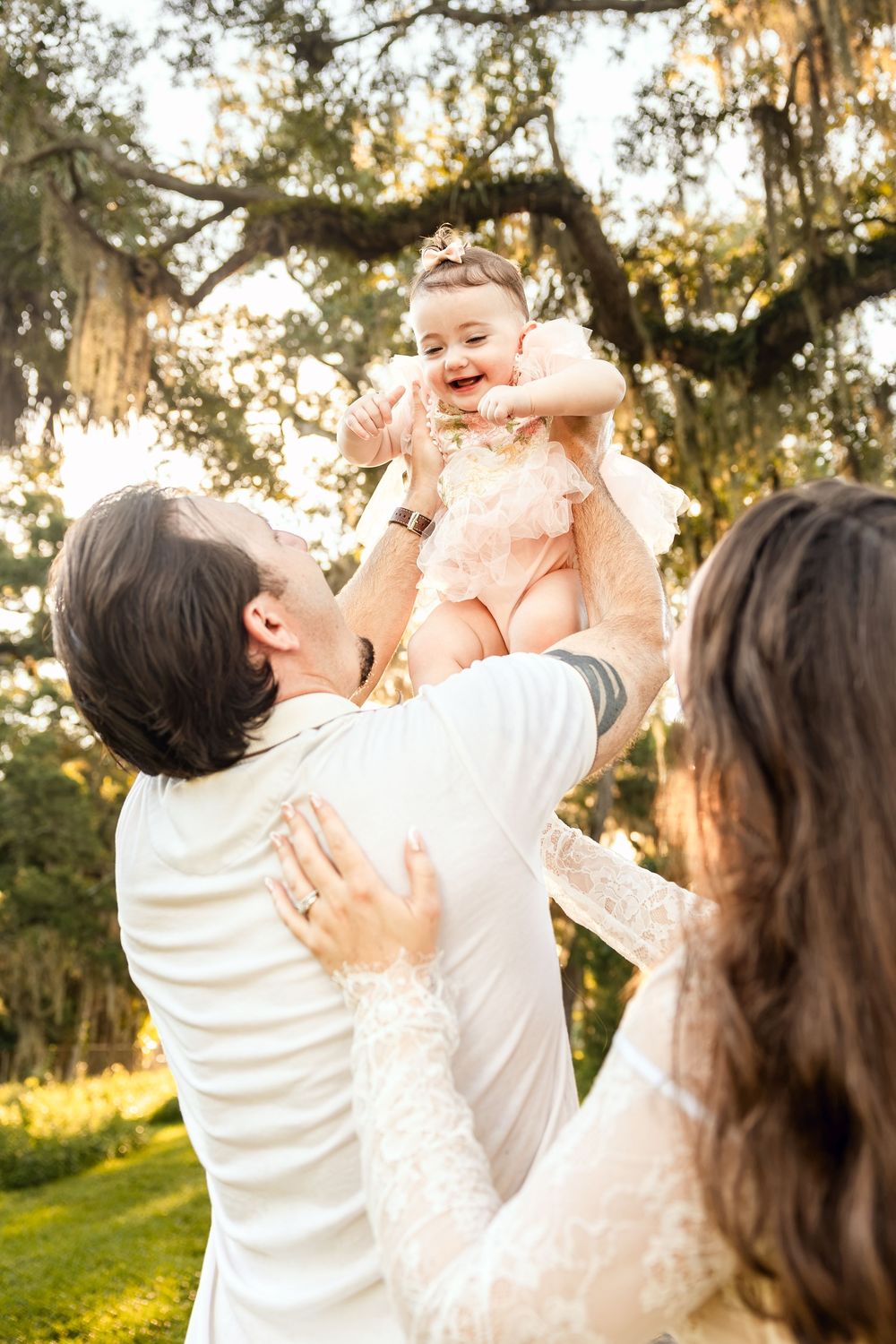 A joyful baby in a pink dress is lifted by her father while mother watches, outdoors under a large oak tree.