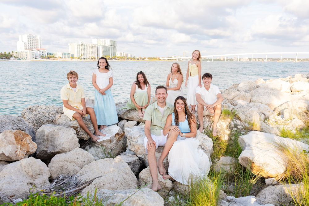 Family portrait on rocky shoreline with city skyline and blue water backdrop on a cloudy day.