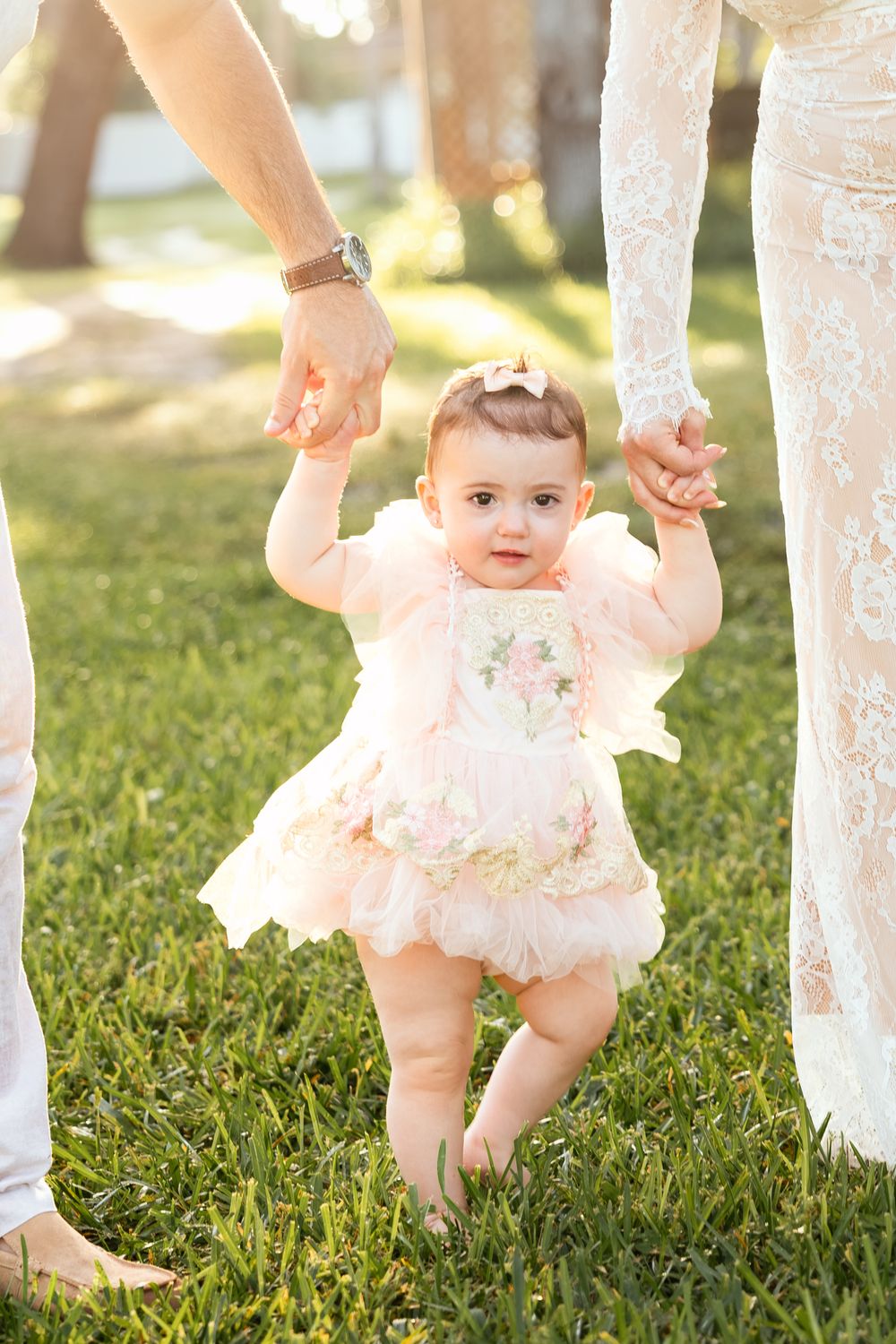 Adorable baby girl in a pink tutu dress learning to walk outdoors, held by parents on green grass.