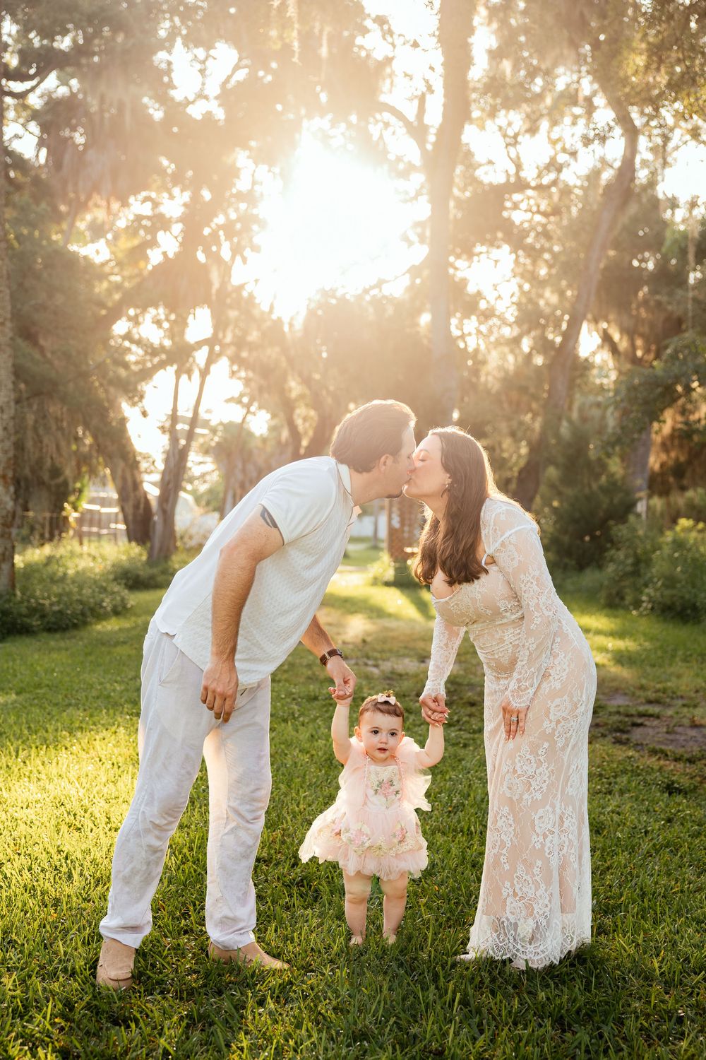 Young couple kissing while holding their baby girl's hands in a sunlit park during golden hour.