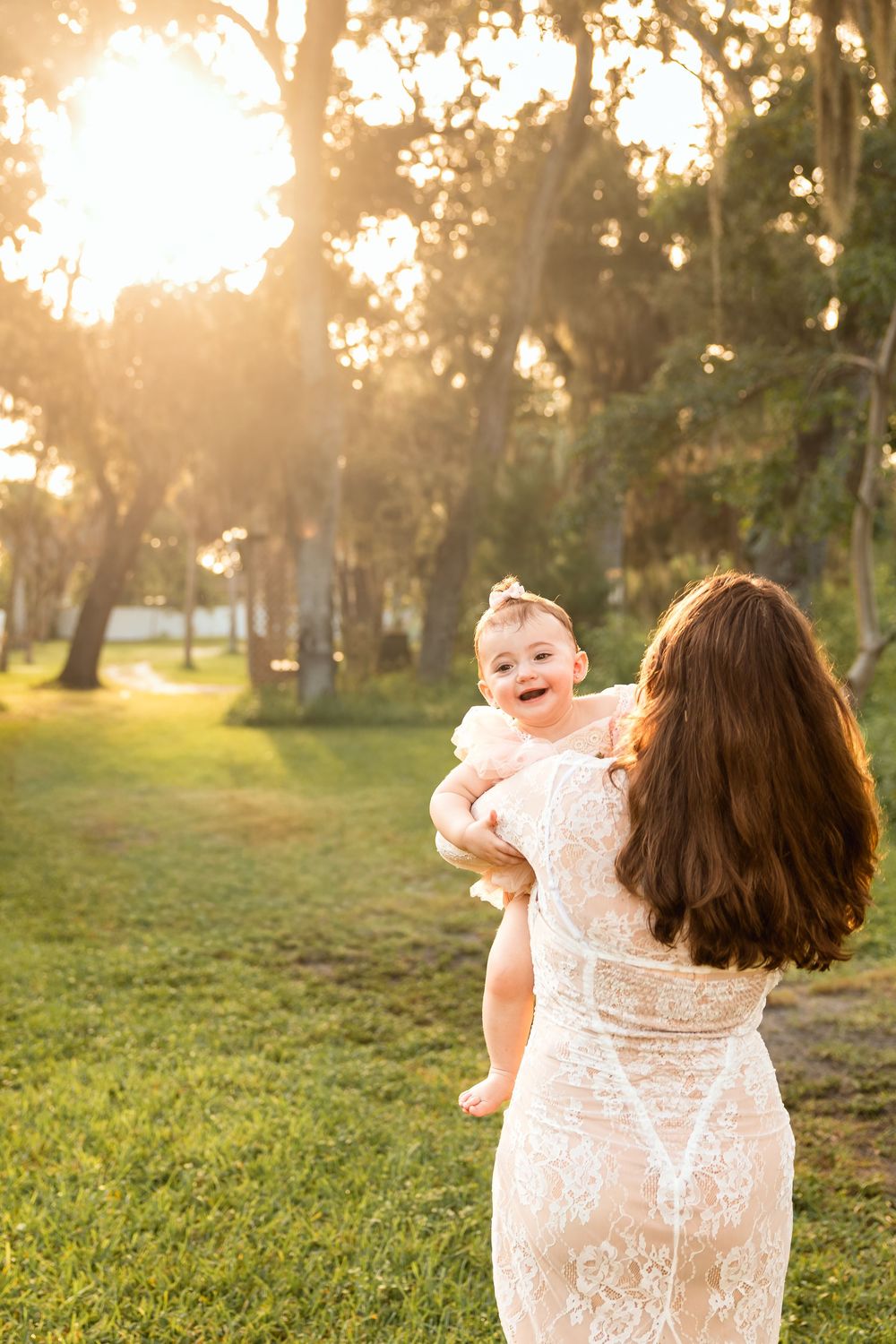 A mother in a lace dress holds her laughing baby girl in a sunlit park during golden hour.