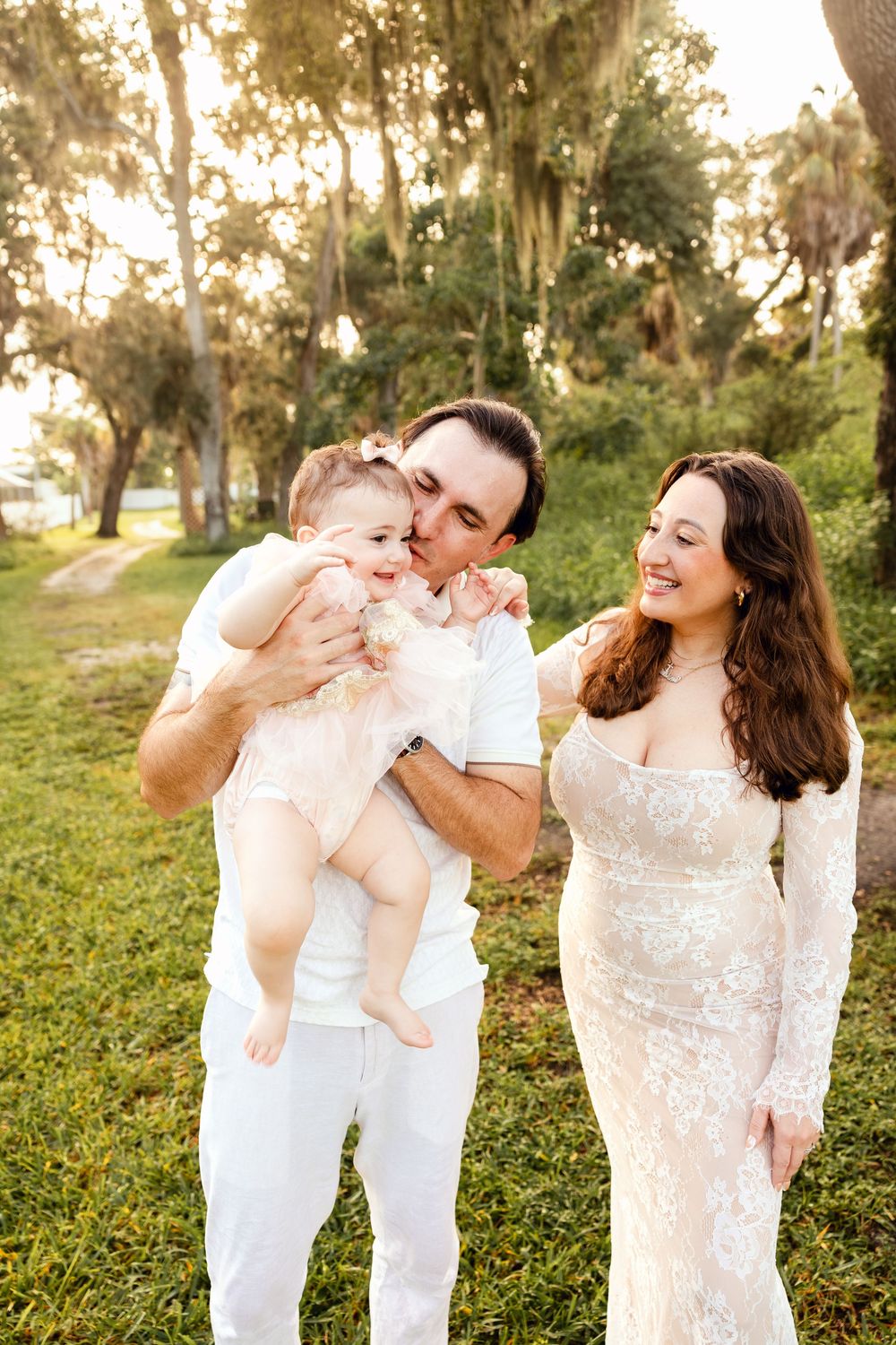 A happy family with a man in white kissing a baby girl while a woman in lace dress smiles outdoors.