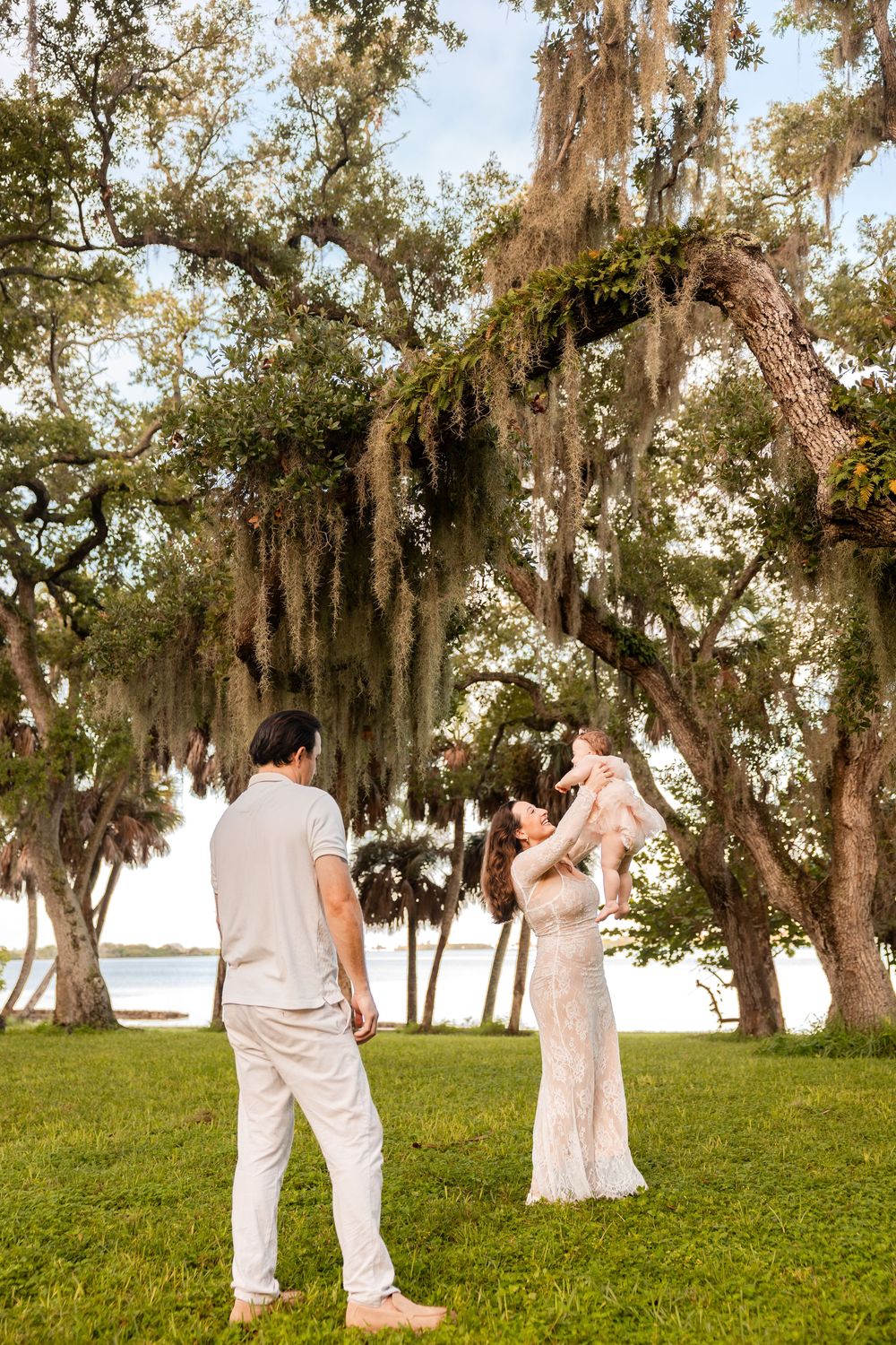 A couple in white outfits lifts their baby under Spanish moss-draped oak trees near a serene lake.