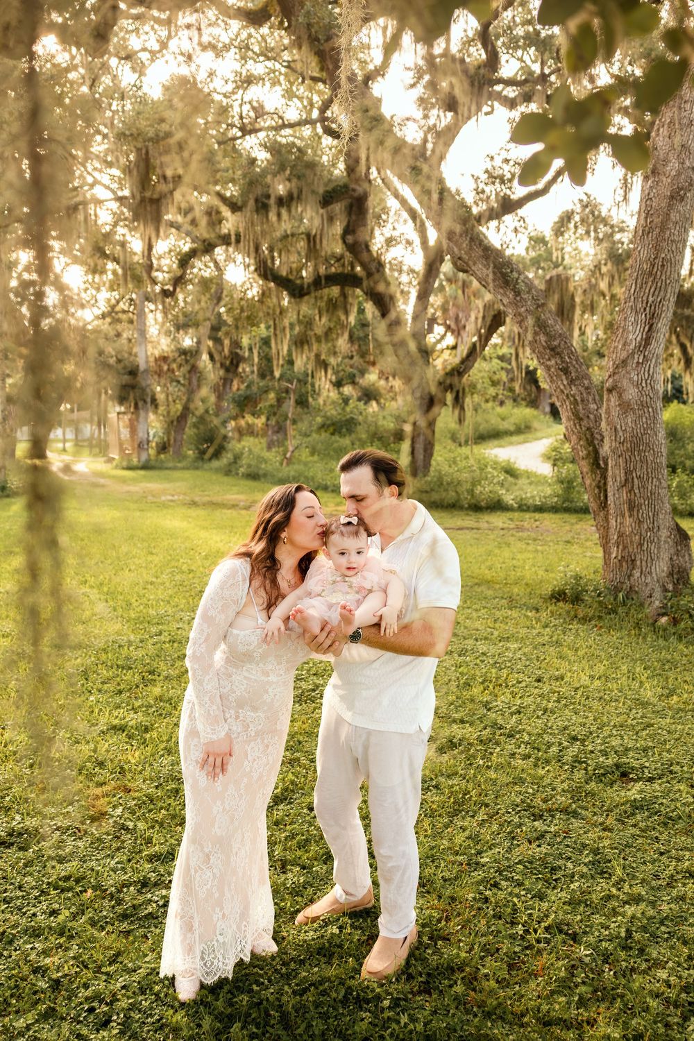 Happy family with parents in white outfits kissing their baby girl under moss-draped oak trees at sunset.