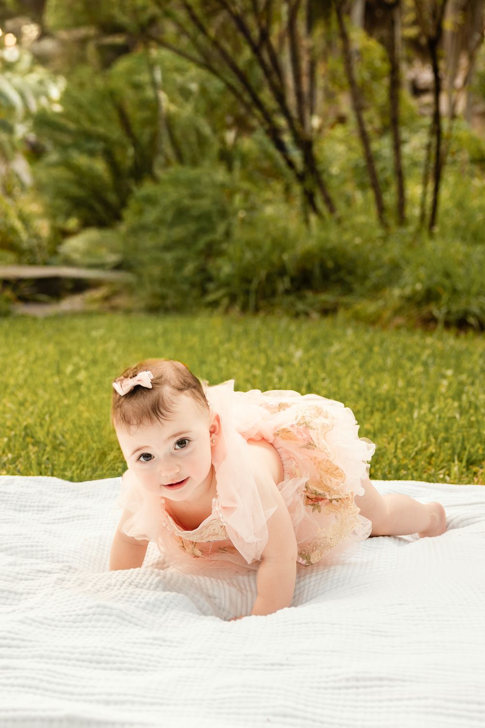 Baby girl in pink tutu dress crawling on white blanket in lush green garden outdoor photo.