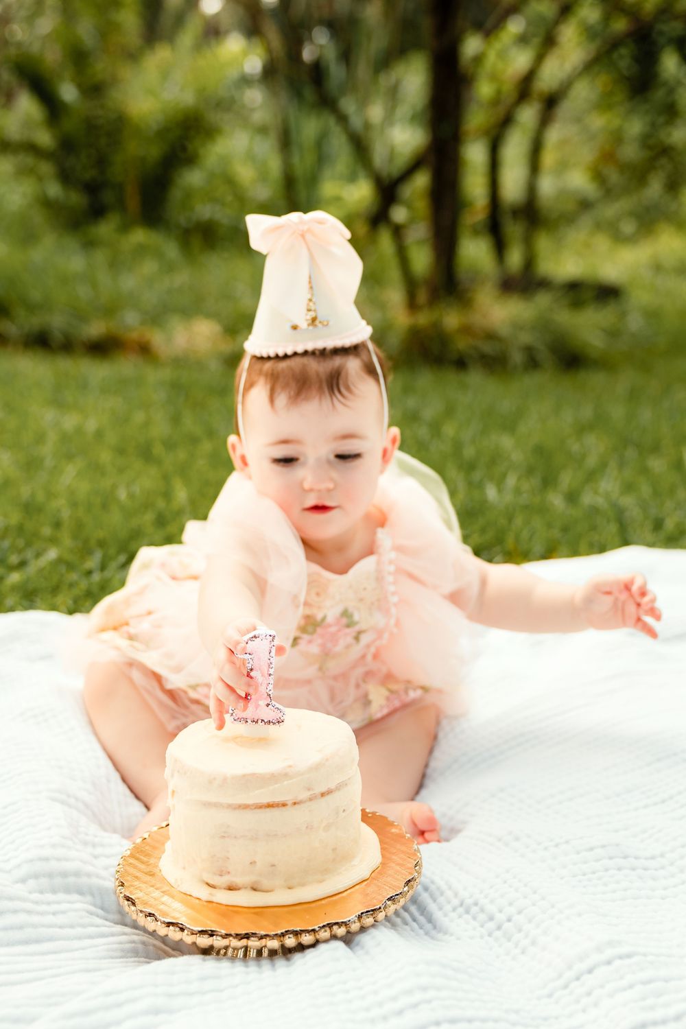 Baby girl in pink floral outfit and party hat reaches for her first birthday smash cake on a blanket outdoors.
