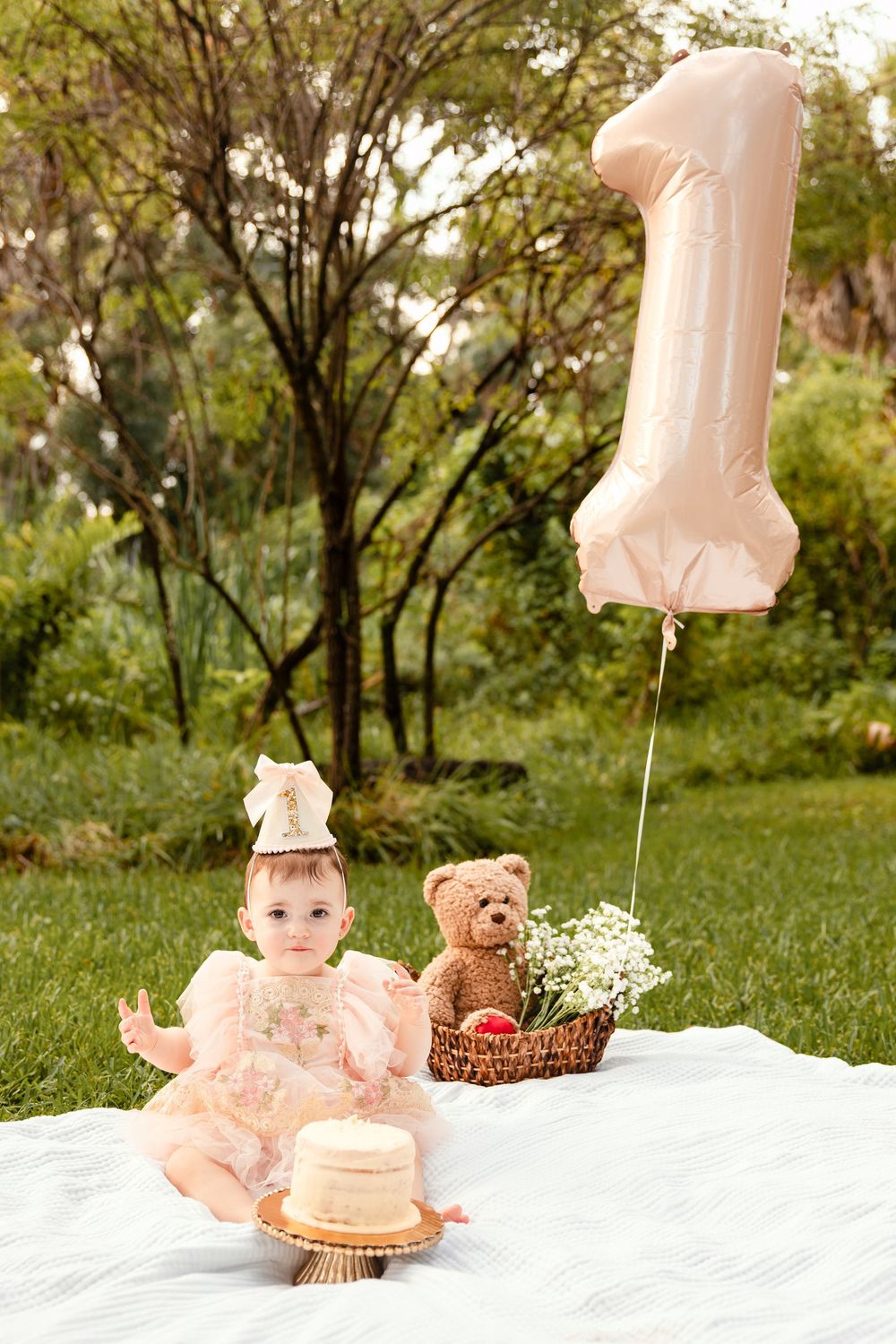 Baby girl in pink dress celebrates 1st birthday with cake, teddy bear, and number one balloon outdoors.