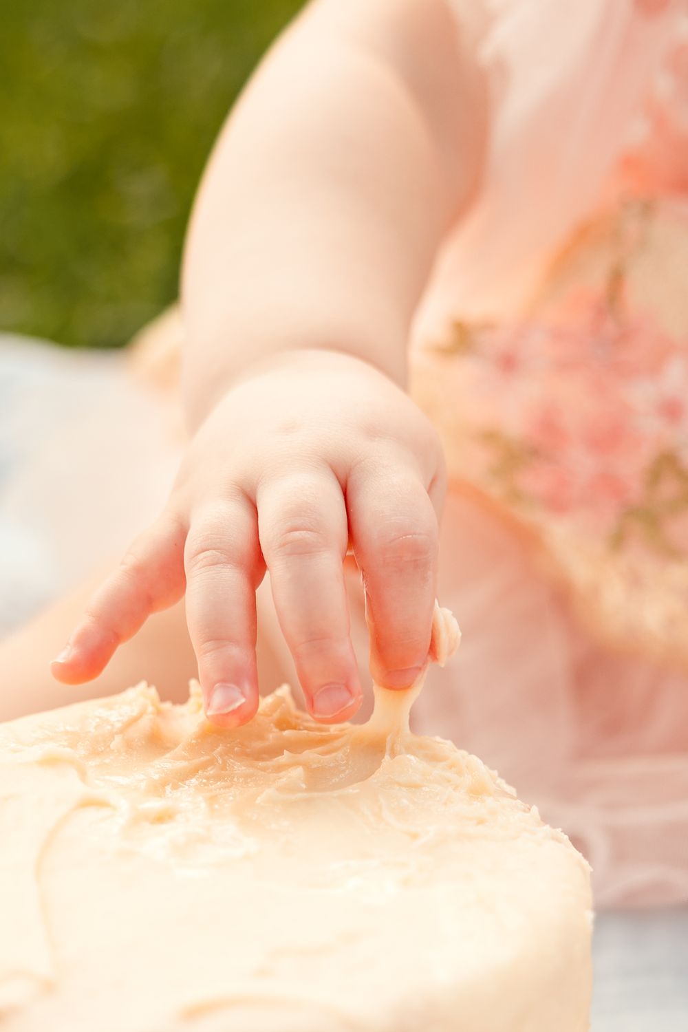 Close-up of a baby's hand touching a cream-frosted smash cake during a first birthday celebration.