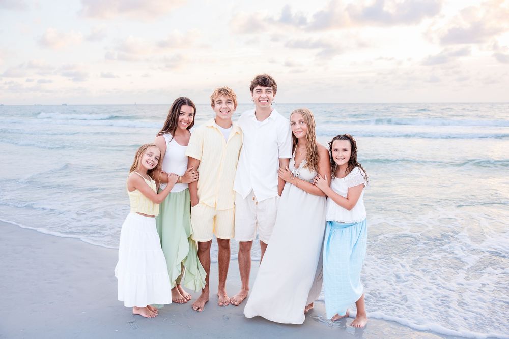Six children smiling together on a sandy beach at sunset wearing coordinated pastel outfits with ocean waves behind them.