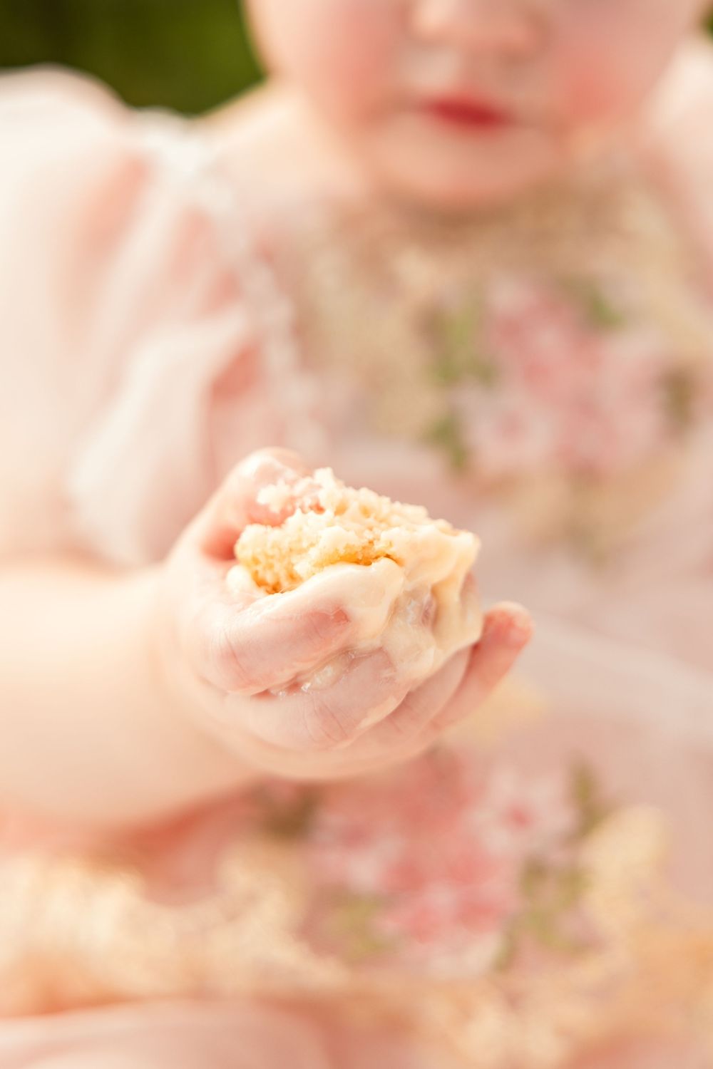 A baby's hand holds a smashed piece of birthday cake during a first birthday cake smash.