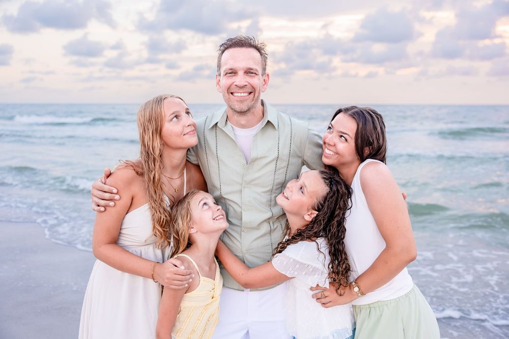 Happy family of five posing together on a sandy beach with ocean waves and pastel sunset sky in the background.