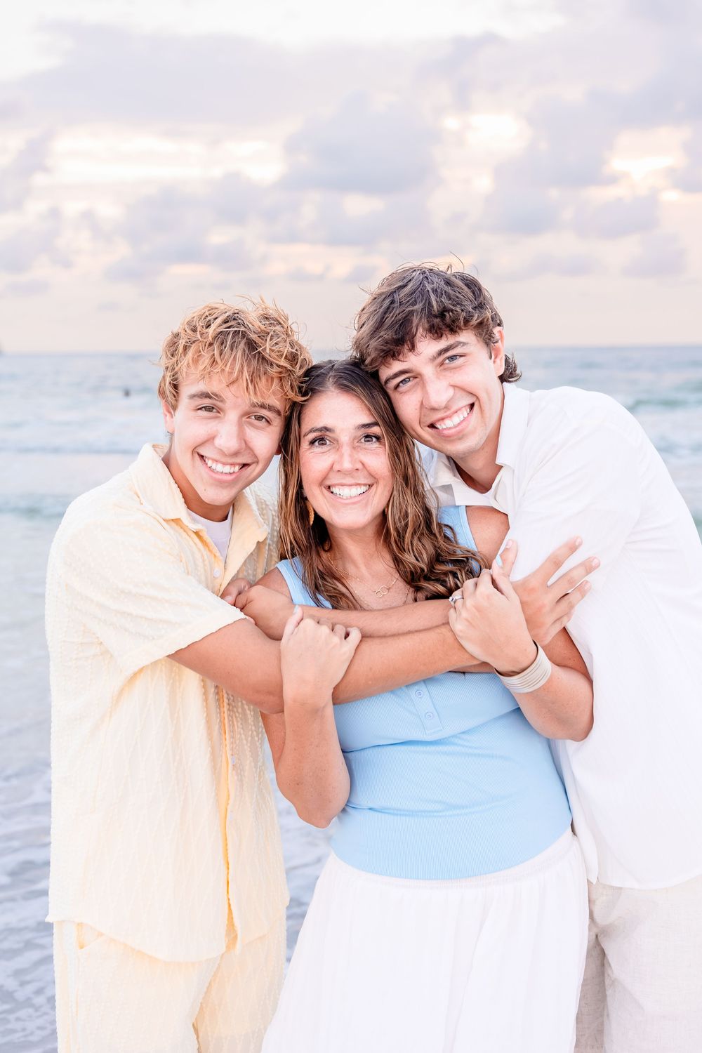 Three smiling young adults posing together on a beach at sunset, two males embracing a female in a blue top.